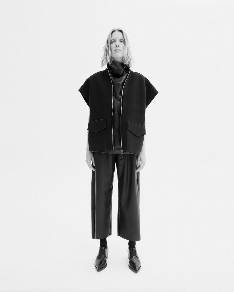 Monochrome fashion shot of a woman in a dark sleeveless jacket, dark top, and culottes posing against a bright white backdrop in a studio.