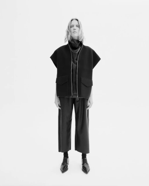 Monochrome fashion shot of a woman in a dark sleeveless jacket, dark top, and culottes posing against a bright white backdrop in a studio.