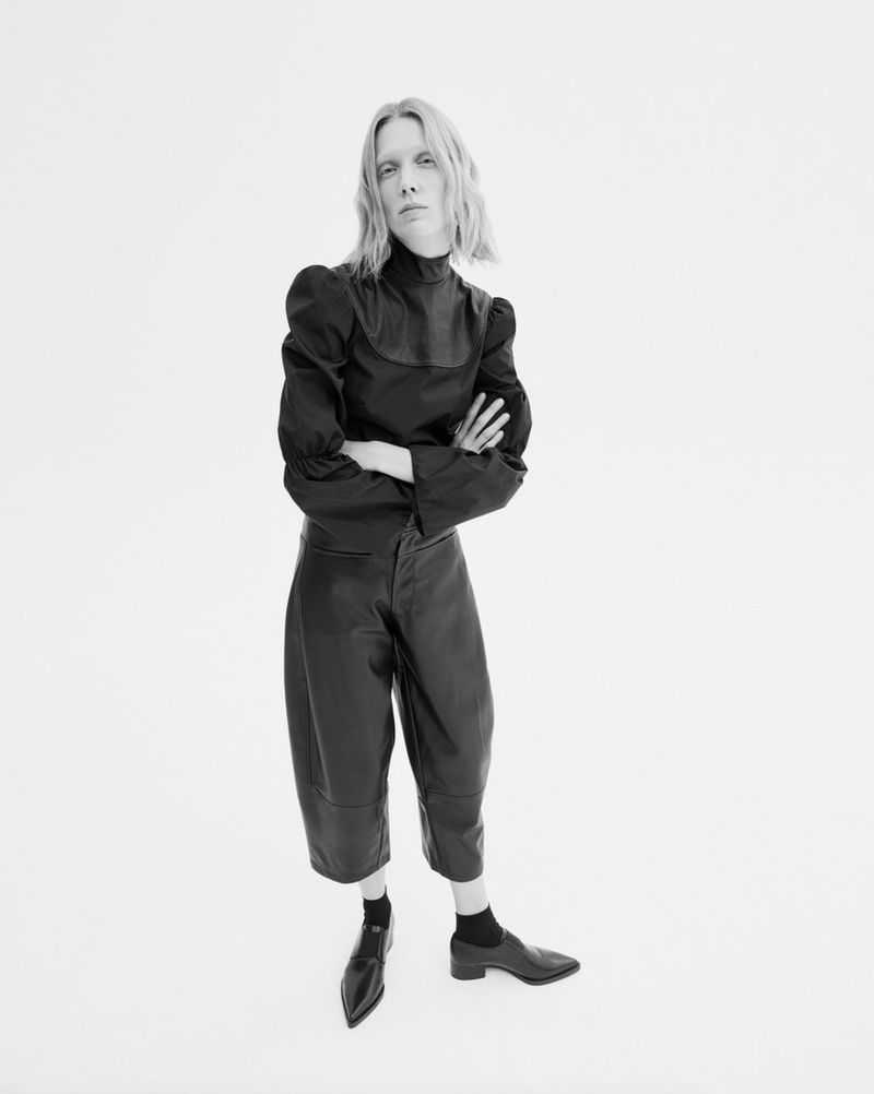 Model Annely Bouma standing with arms crossed wearing a black high-neck puff-sleeve top and leather culottes against a white background.