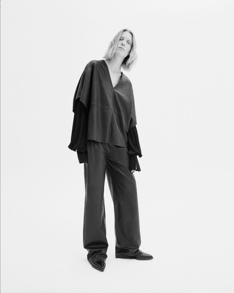 Fashionable adult female poses in a black top and wide leg pants against a white backdrop, showcasing a minimalist aesthetic.