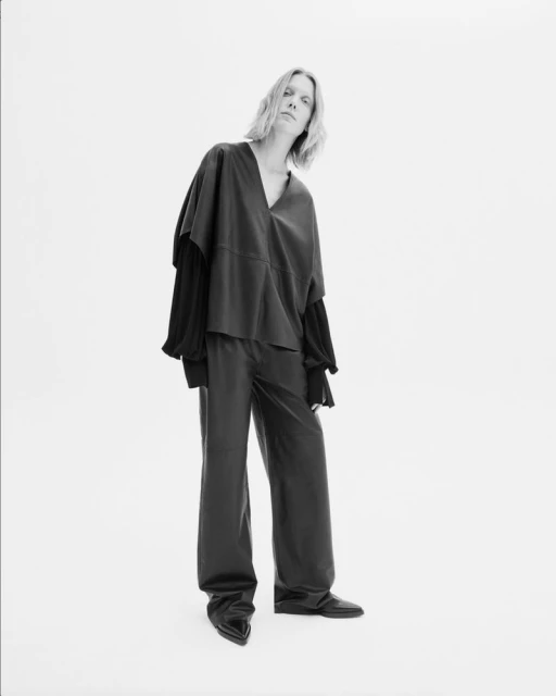 Fashionable adult female poses in a black top and wide leg pants against a white backdrop, showcasing a minimalist aesthetic.