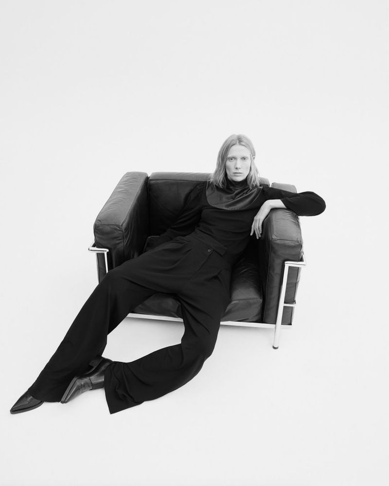 Black and white portrait of a woman sitting in a leather chair in a photo studio, wearing a black turtleneck sweater and wide-leg pants.