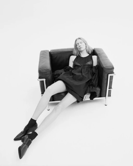 Woman poses in a black dress and black shoes, seated in a black chair in a bright photo studio. The scene is captured in monochrome.