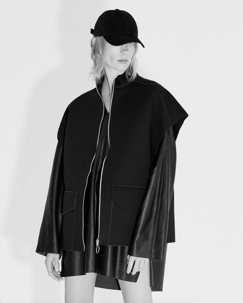 Studio shot of a slender adult woman wearing a black baseball cap, a layered outfit of leather and fabric, against a white backdrop.