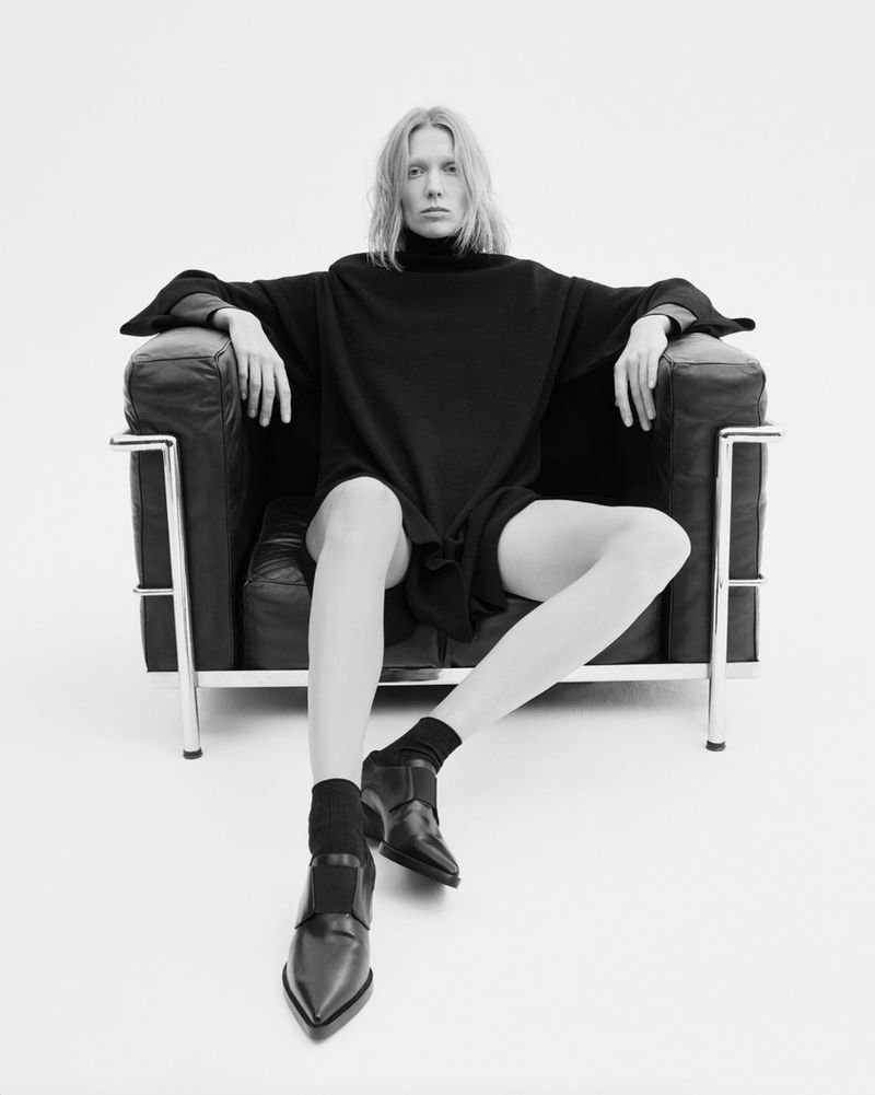 Blonde adult female poses in a black turtleneck dress, socks, and loafers, seated in a black leather Le Corbusier chair against a white background.