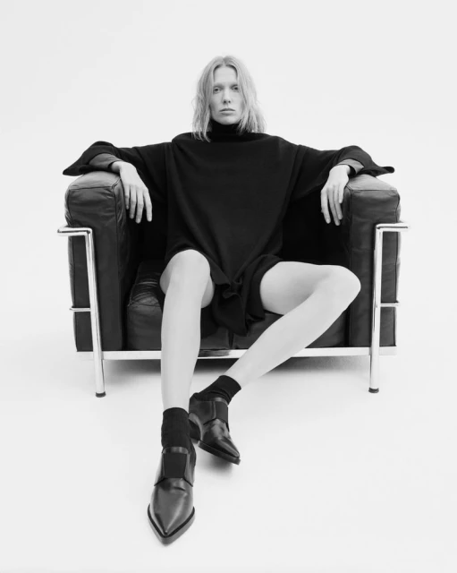 Blonde adult female poses in a black turtleneck dress, socks, and loafers, seated in a black leather Le Corbusier chair against a white background.