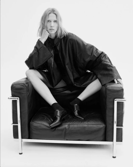Black and white portrait of a fair-skinned woman with long blonde hair posing in a leather chair against a clean white background.