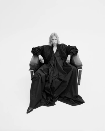 Model Annely Bouma seated in a black leather armchair wearing an oversized black jacket and draped floor-length black gown on white seamless.