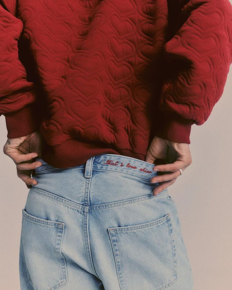 Close-up of a person wearing a red sweater with heart patterns and light blue jeans with red embroidered text on the waistband.