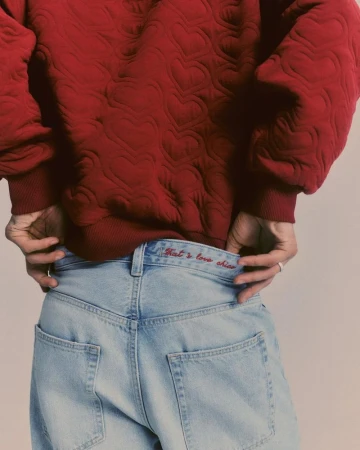 Close-up of a person wearing a red sweater with heart patterns and light blue jeans with red embroidered text on the waistband.