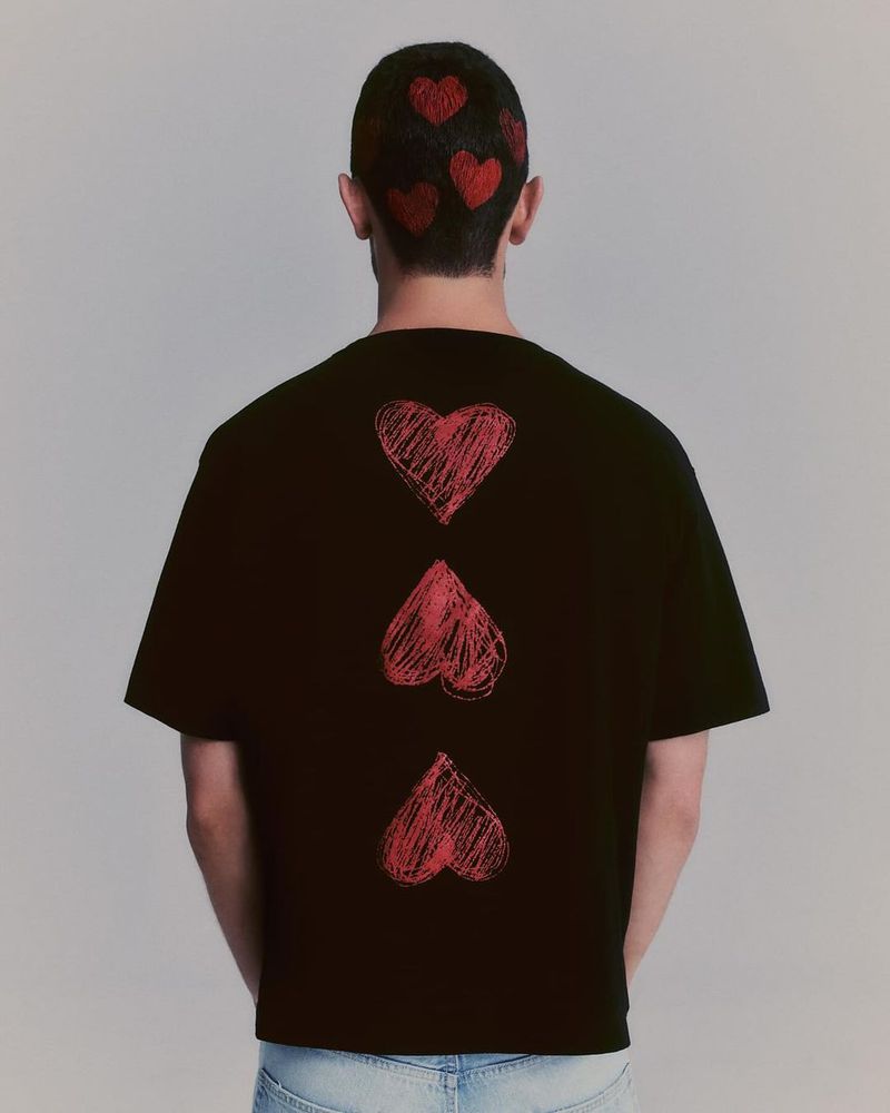 Person wearing a black t-shirt with red heart illustrations on the back and head against a light gray studio backdrop.