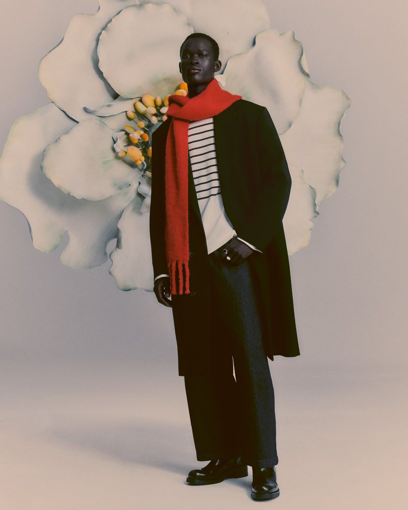 Full-length portrait of a Black male model in a black coat and red scarf standing in front of a large flower backdrop in a studio.