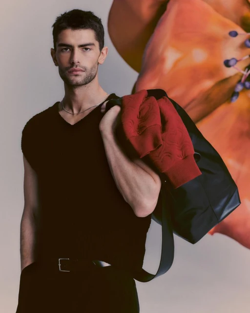Portrait of a handsome adult man in a black top and pants, carrying a black duffel bag and a red jacket, with a floral background.