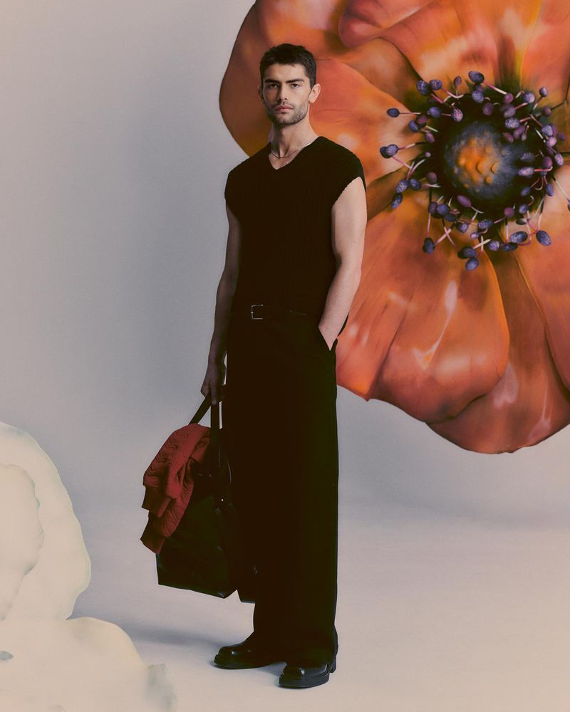 A brunette man in black clothing and black dress shoes poses with a duffel bag against a pale studio backdrop with an oversized flower prop.