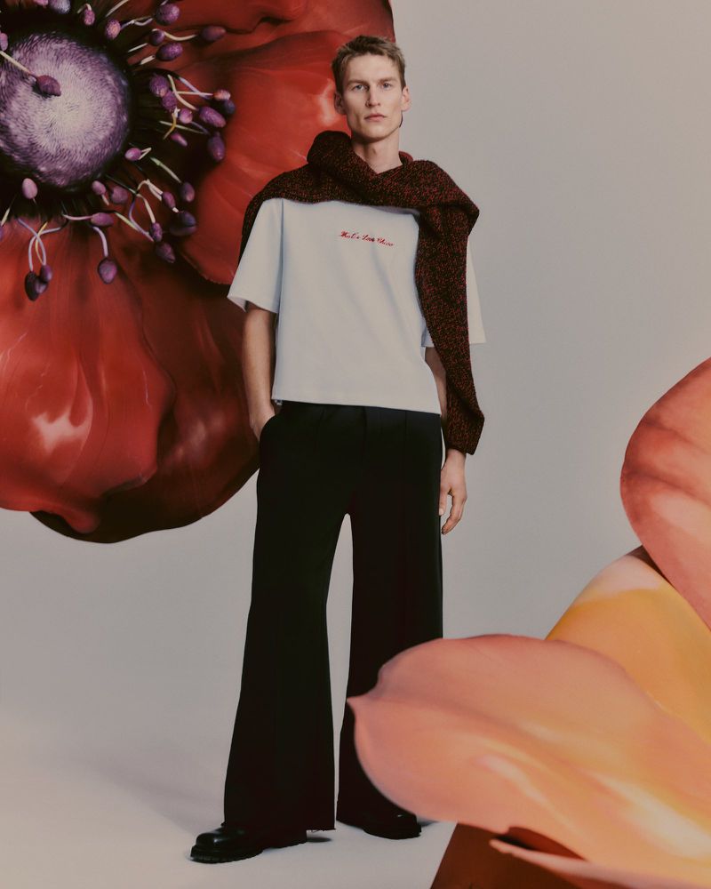 A full-length shot showcases a stylish adult male model in a casual white T-shirt and flared black pants, accessorized with a draped scarf and set against a flower backdrop.