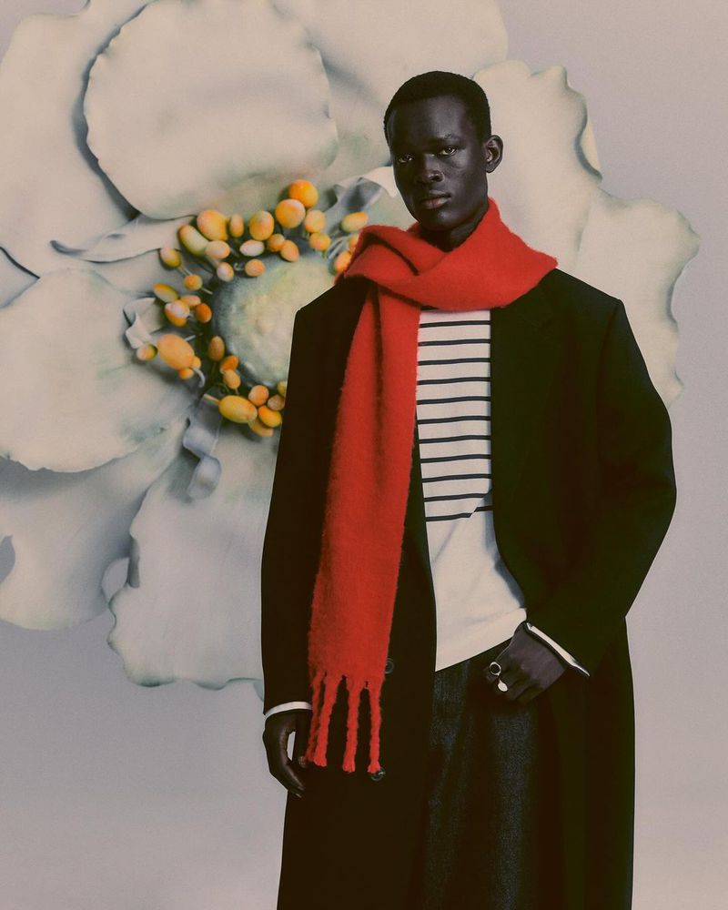 Fashionable portrait of a dark-skinned man in a black coat and red scarf against a flower backdrop, captured in a studio setting.