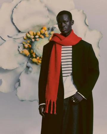 Fashionable portrait of a dark-skinned man in a black coat and red scarf against a flower backdrop, captured in a studio setting.