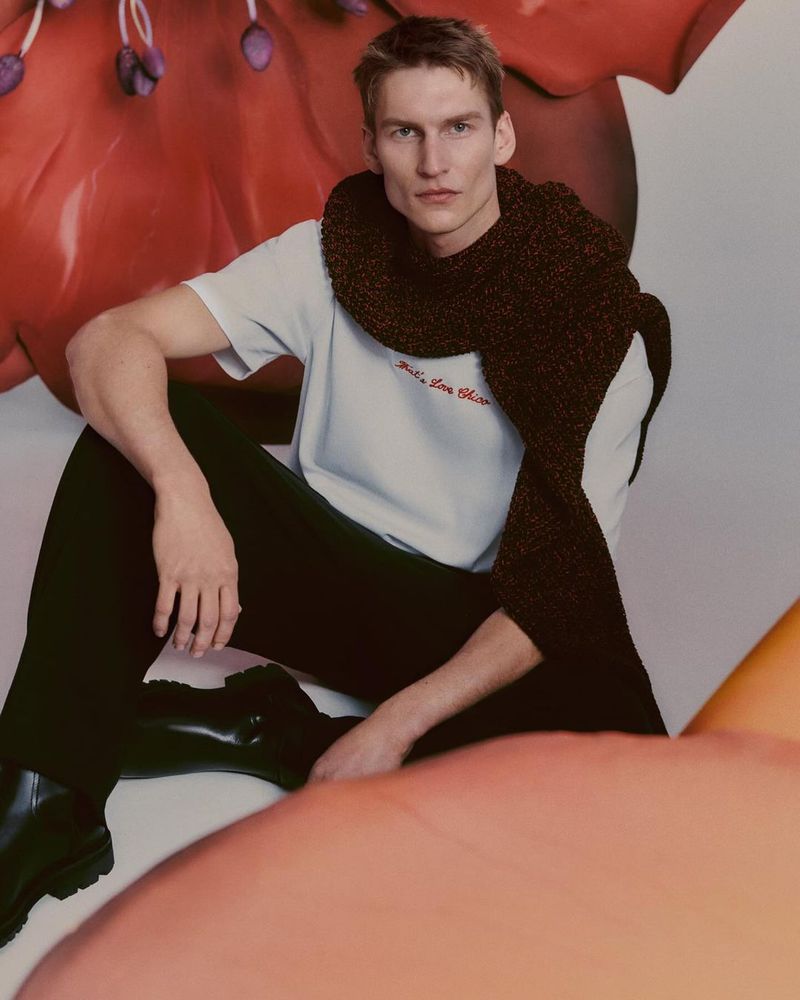 A blonde male model poses indoors wearing a light-blue t-shirt, black trousers, and dark-brown scarf. He is seated in front of a floral background.
