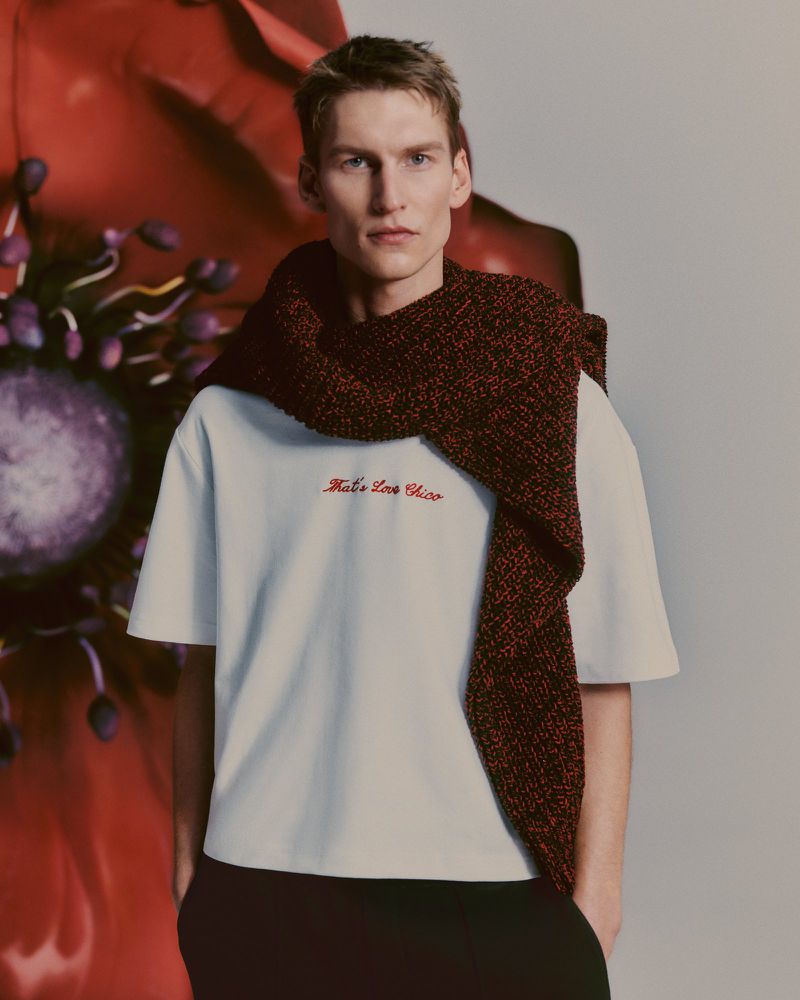 Male fashion model poses indoors wearing a white t-shirt and red knit scarf against a blurred red floral background.