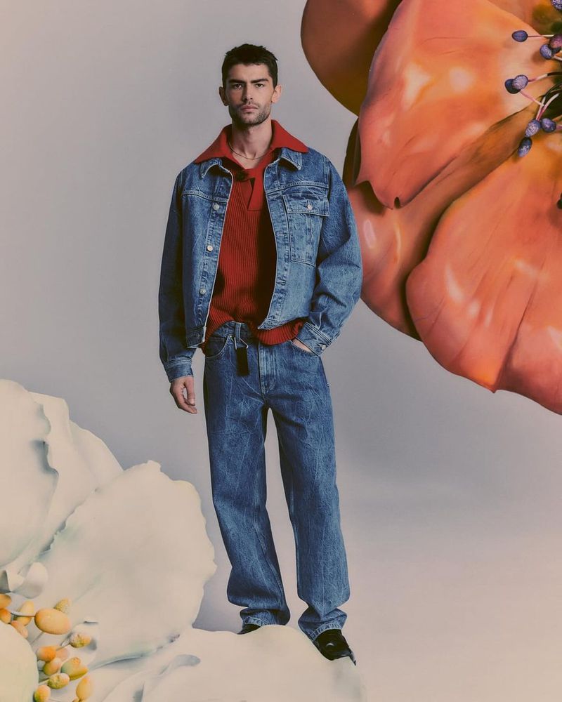 A full-length shot showcases a male model in a denim jacket and jeans against a studio backdrop with a floral art installation.