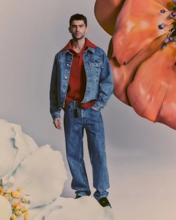 A male model poses in a denim jacket and jeans, standing on a cloud-like surface against a soft grey backdrop with floral accents.
