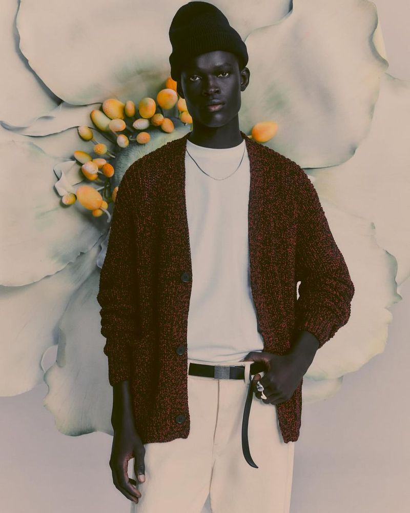 Man in cardigan and white pants stands against flower backdrop, wearing a beanie hat and silver chain. Indoor fashion shot.