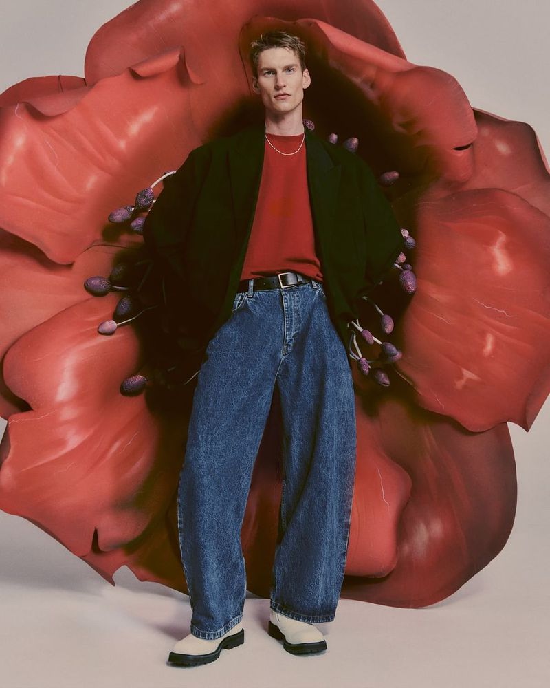 Male fashion model poses in oversized denim jeans and a red shirt with a black jacket against a giant red flower backdrop.
