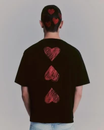 Rear view of a man in a black t-shirt featuring three red sketched hearts and red heart hair art against a neutral studio backdrop.