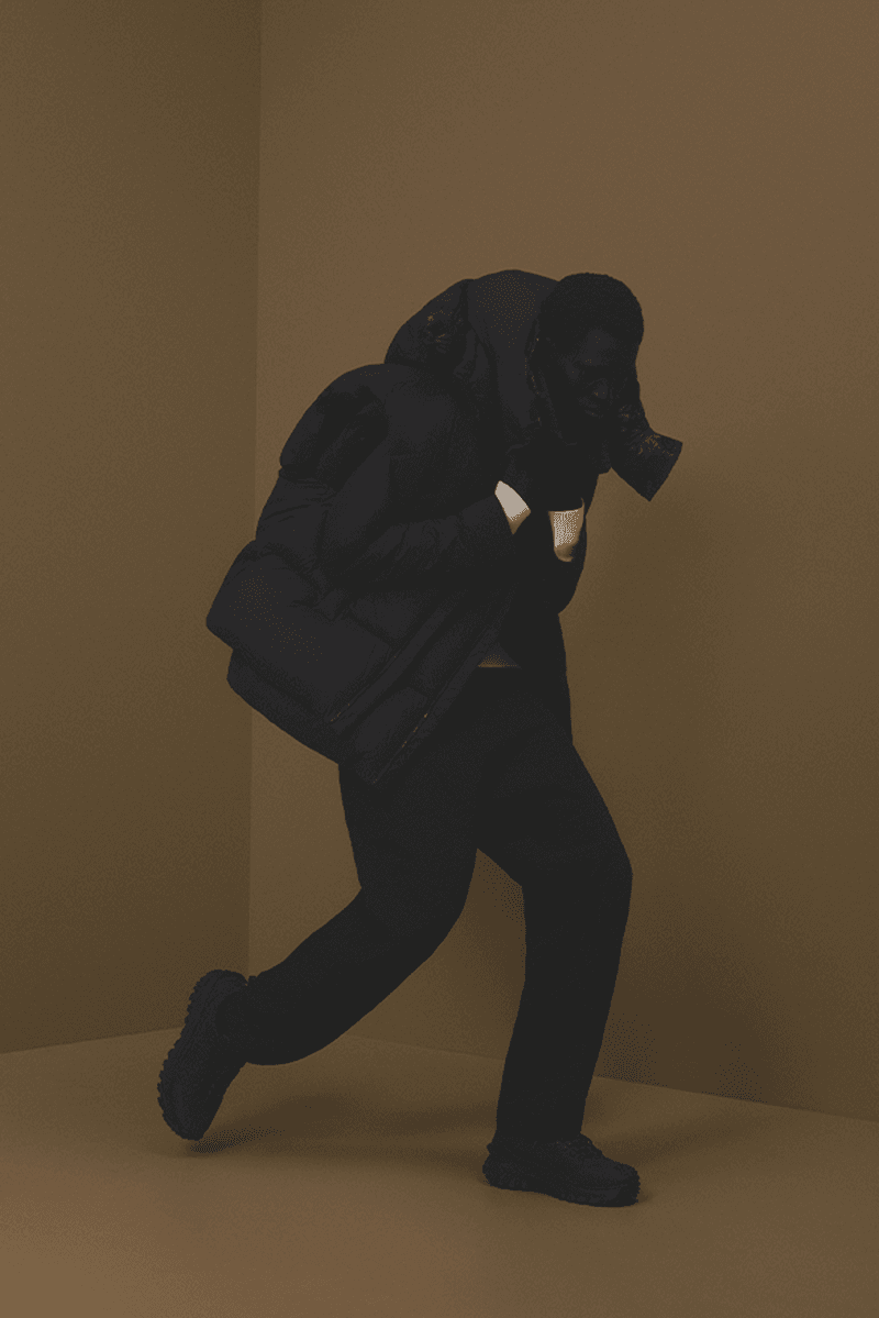 Person in black clothing and a gas mask walking against a brown background in a photo studio environment. Dark color palette.