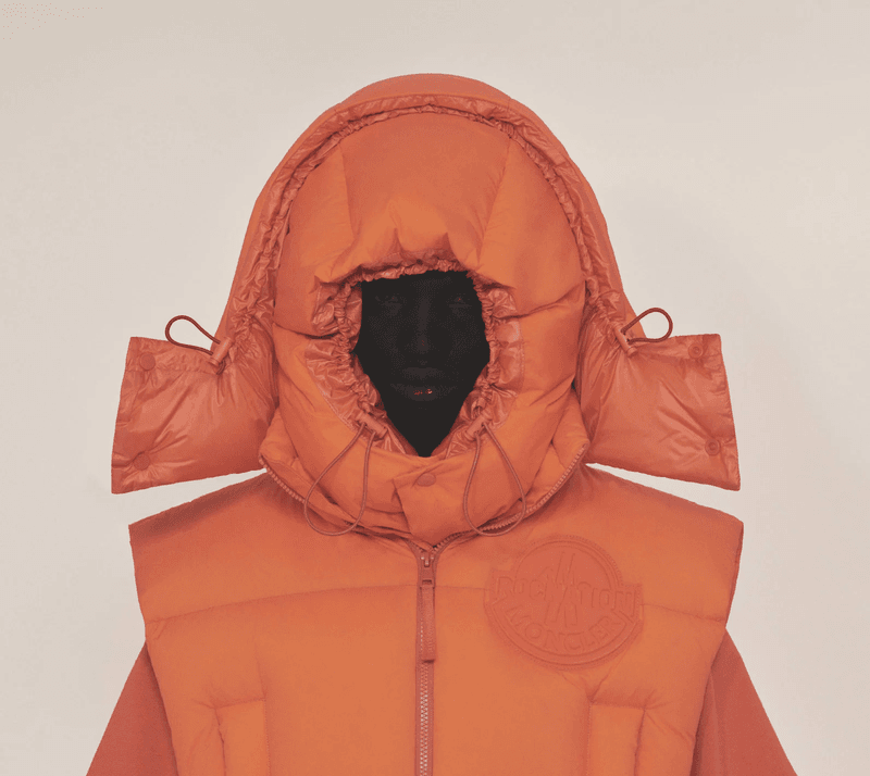 Person wearing an orange hooded puffer vest. The puffer vest features an emblem. The background is a light neutral color.