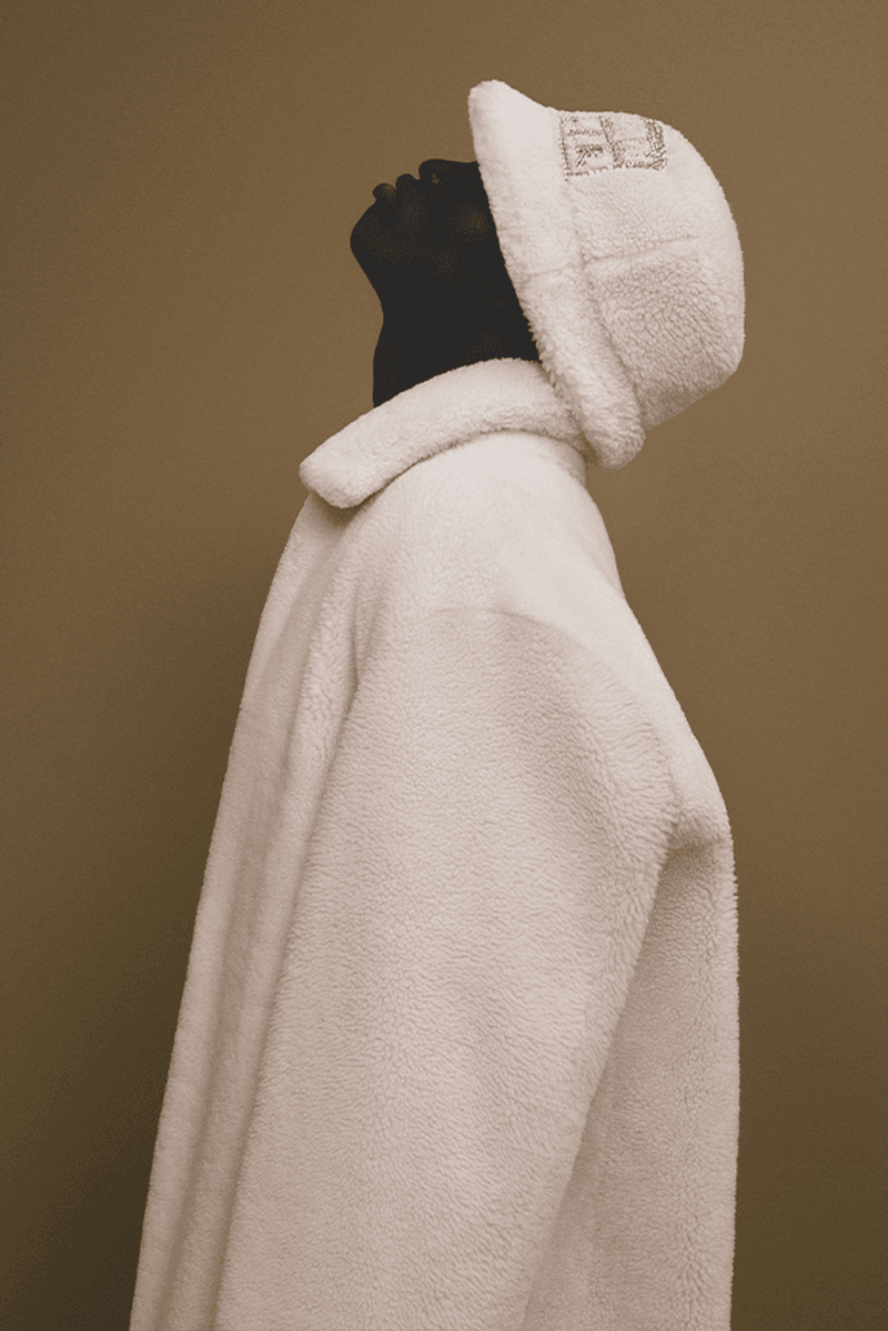 Profile shot of an adult fashion model with dark skin wearing a textured white sherpa-style bucket hat and coat on a neutral backdrop.