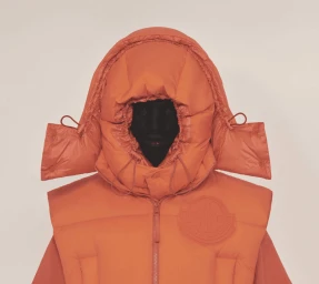 Model wearing an orange padded puffer vest with an oversized hooded collar against a neutral background