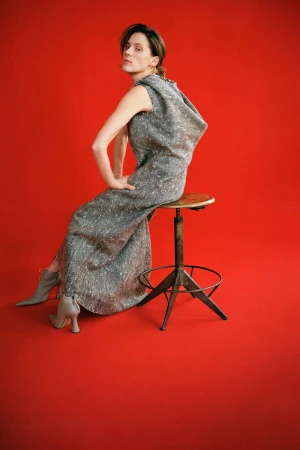Model seated on a studio stool wearing a sleeveless grey draped gown and pointed heeled ankle boots against a vibrant red backdrop.
