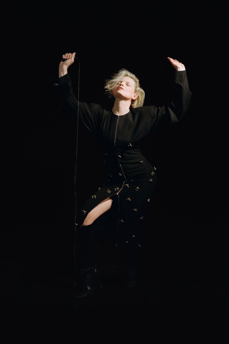 Woman in black dress with blonde hair poses in dark studio with arms raised while holding black cable with silver attachments.