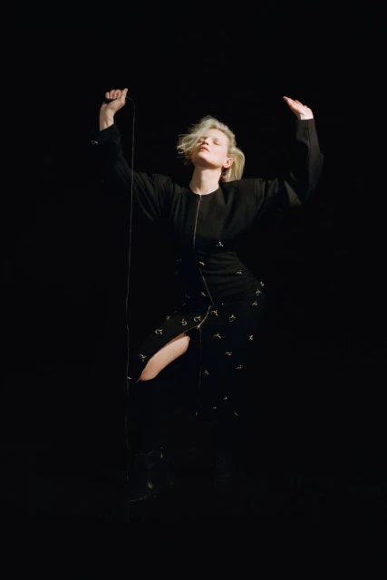 Woman in black dress with blonde hair poses in dark studio with arms raised while holding black cable with silver attachments.