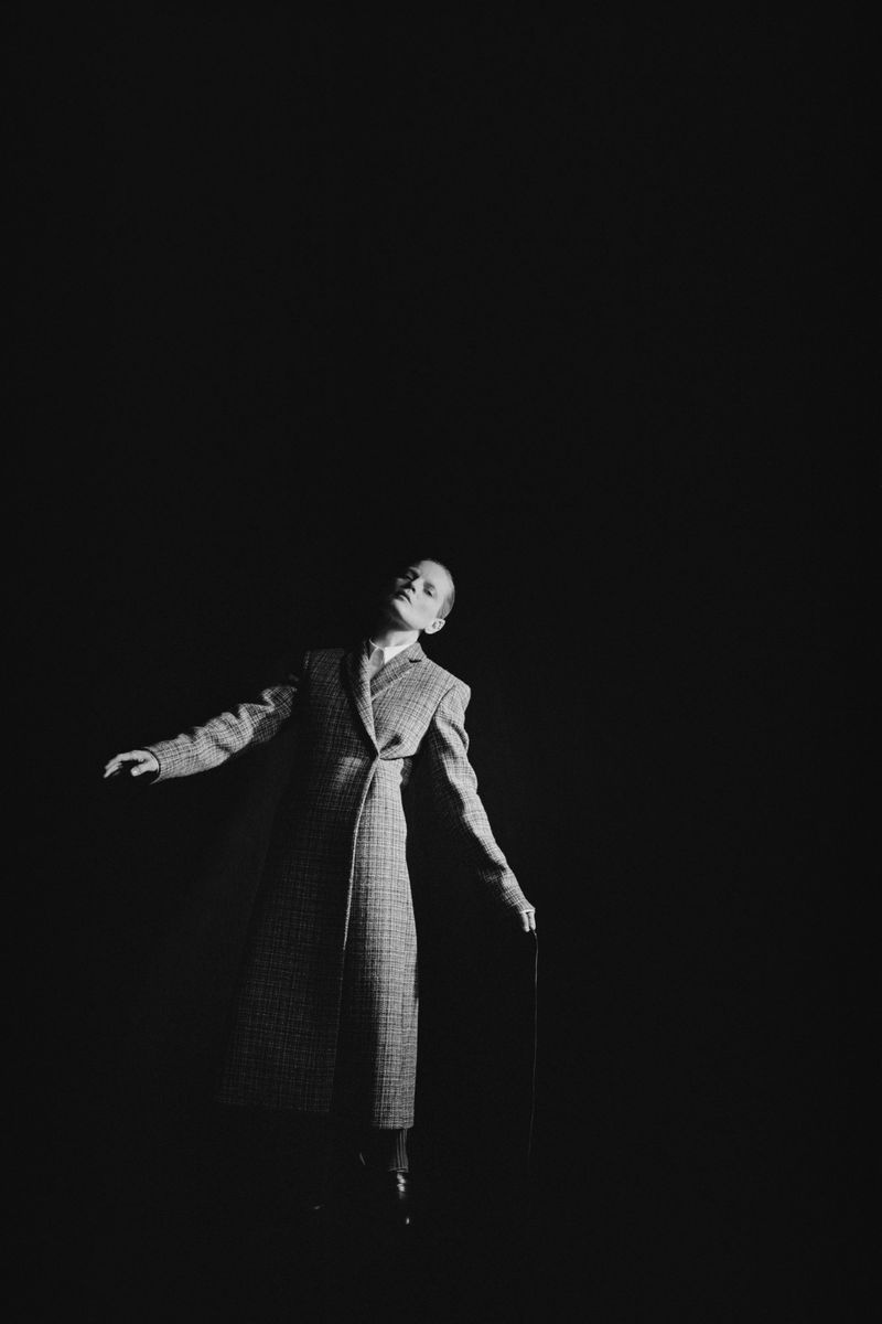 Monochrome portrait of a male fashion model wearing a patterned overcoat and holding a cane, set against a stark black background.