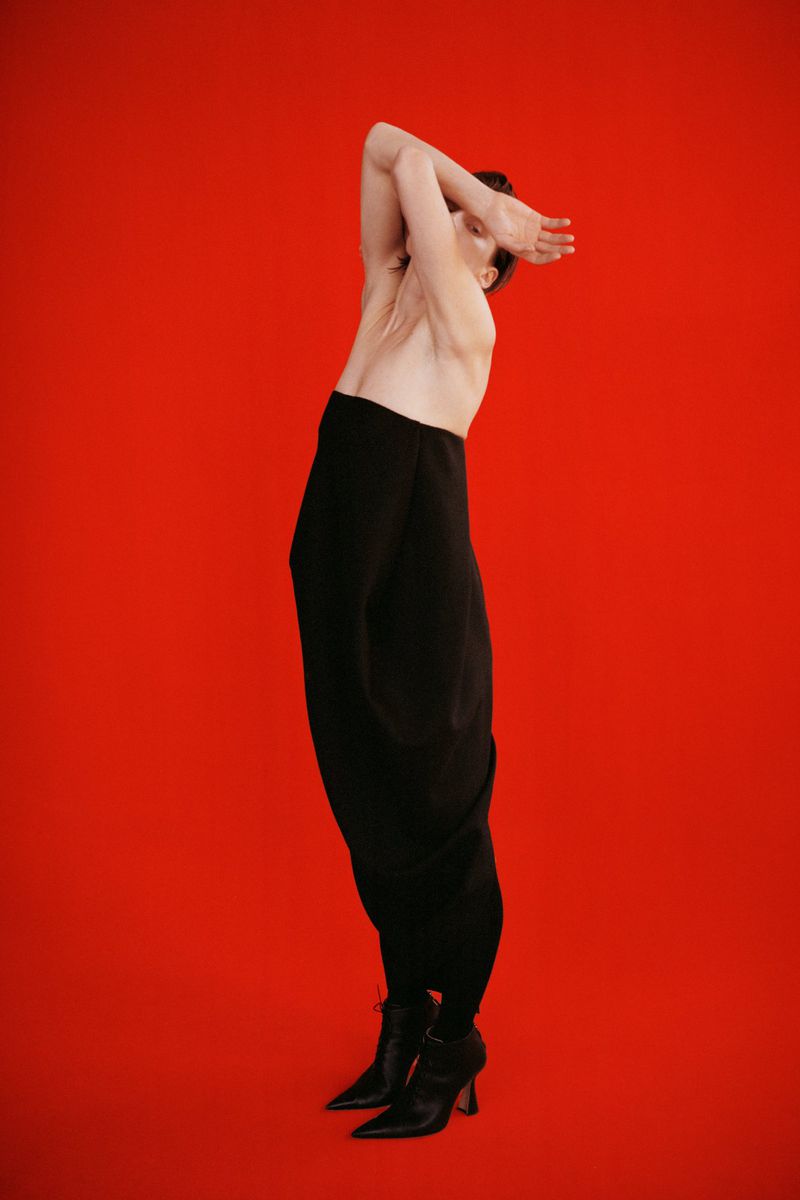 Fashion photo of a person with arms raised posing against a red backdrop, wearing loose black pants and heeled boots in a studio setting.