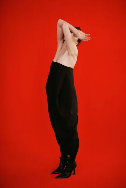 Fashion photo of a person with arms raised posing against a red backdrop, wearing loose black pants and heeled boots in a studio setting.