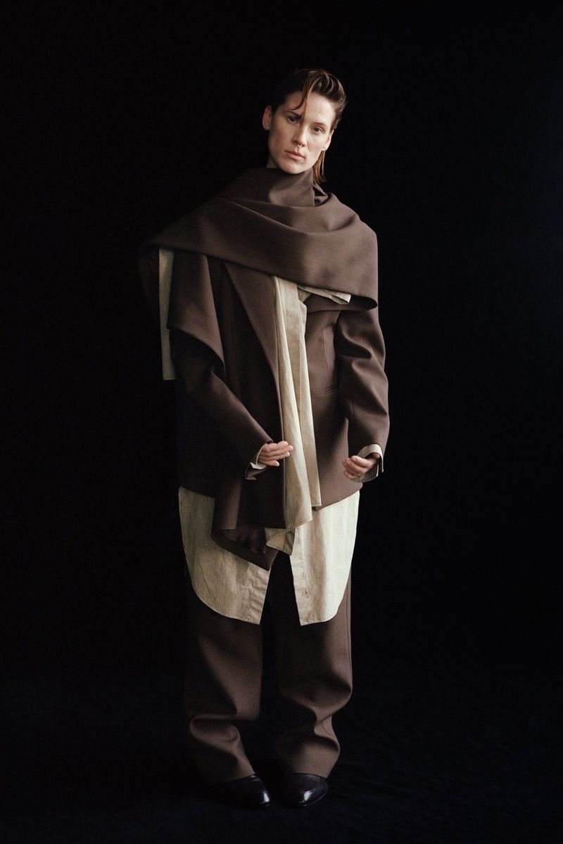 Fashionable adult in brown pantsuit and draped brown scarf against a black backdrop in a full portrait. Elegant attire.