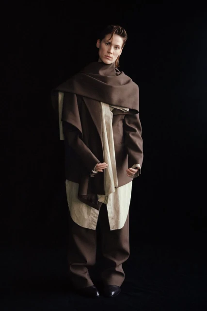 Fashionable adult in brown pantsuit and draped brown scarf against a black backdrop in a full portrait. Elegant attire.