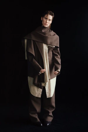 Fashionable adult in brown pantsuit and draped brown scarf against a black backdrop in a full portrait. Elegant attire.