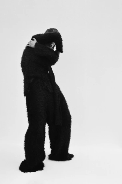 Monochromatic portrait of a slender adult in black fuzzy clothing with dramatic pose against a bright, stark white backdrop in studio.