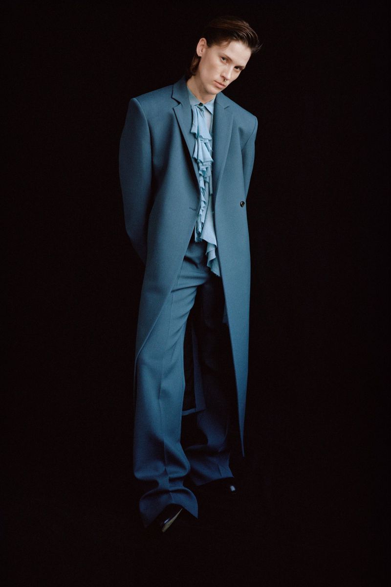 Man in an elegant blue suit with a ruffled shirt standing against a black background in a fashion editorial portrait.