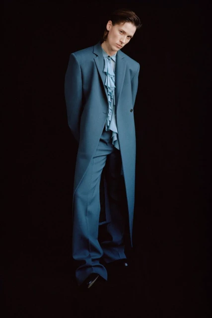 Man in an elegant blue suit with a ruffled shirt standing against a black background in a fashion editorial portrait.