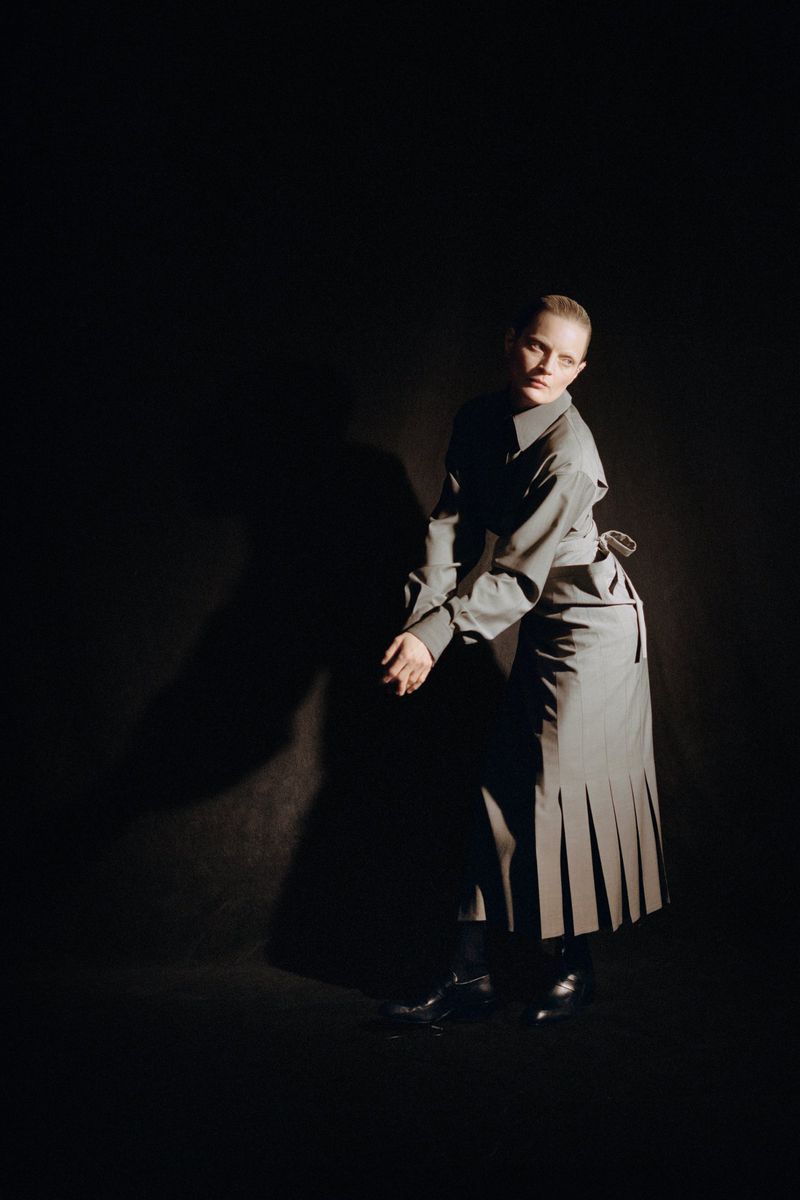 Young woman with fair skin, wearing a grey shirt and pleated skirt, poses in a photo studio with dramatic lighting against a dark backdrop.