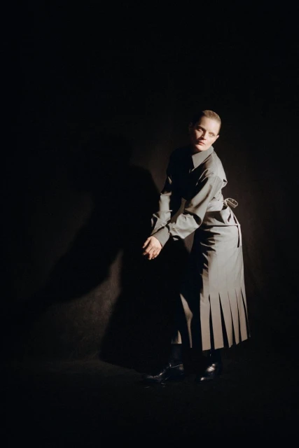 Young woman with fair skin, wearing a grey shirt and pleated skirt, poses in a photo studio with dramatic lighting against a dark backdrop.