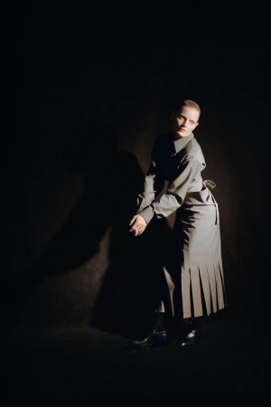Young woman with fair skin, wearing a grey shirt and pleated skirt, poses in a photo studio with dramatic lighting against a dark backdrop.