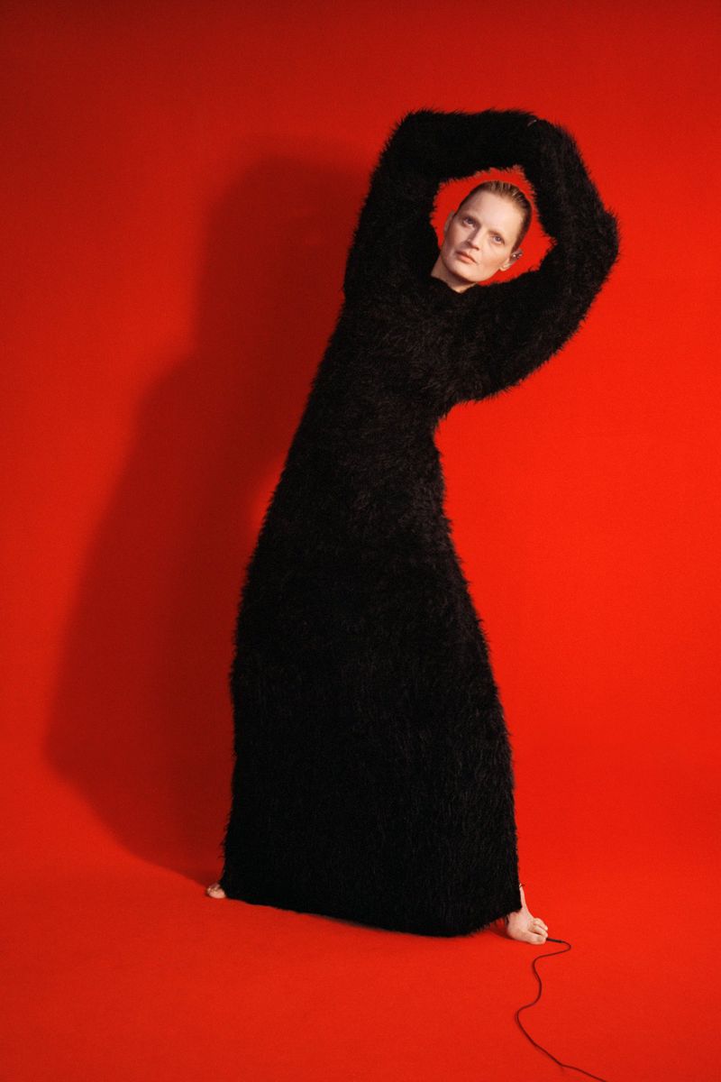 A woman in a floor-length, black, textured gown stands against a vibrant red backdrop in a studio, with her arms raised above her head.