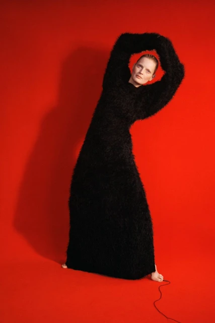 A woman in a floor-length, black, textured gown stands against a vibrant red backdrop in a studio, with her arms raised above her head.