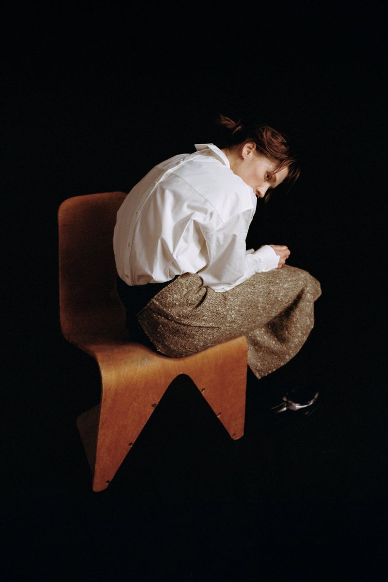 Woman in white shirt and tweed skirt sits sideways on light wood chair in dark, soft studio lighting. Modern portrait photography.